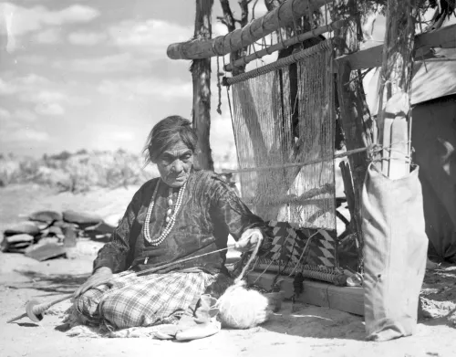 Navajo weaver spinning wool into yarn; full- length, seated, with a loom behind her, Torre'on, New Mexico #126